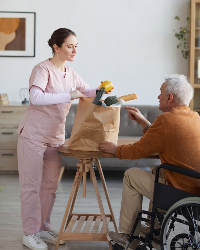 vertical-full-length-portrait-young-female-caregiver-bringing-groceries-senior-man-wheelchair-assistance-food-delivery-concept
